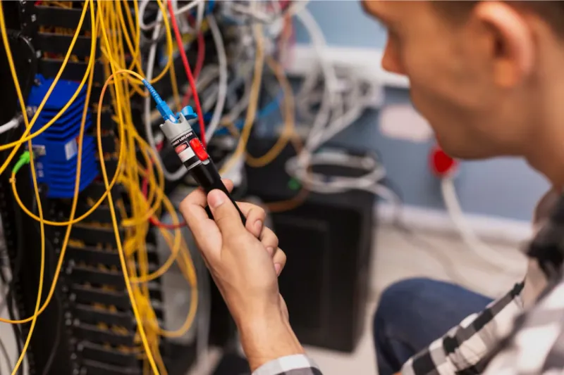 Técnico testando cabos de fibra óptica com uma caneta de luz em um rack.